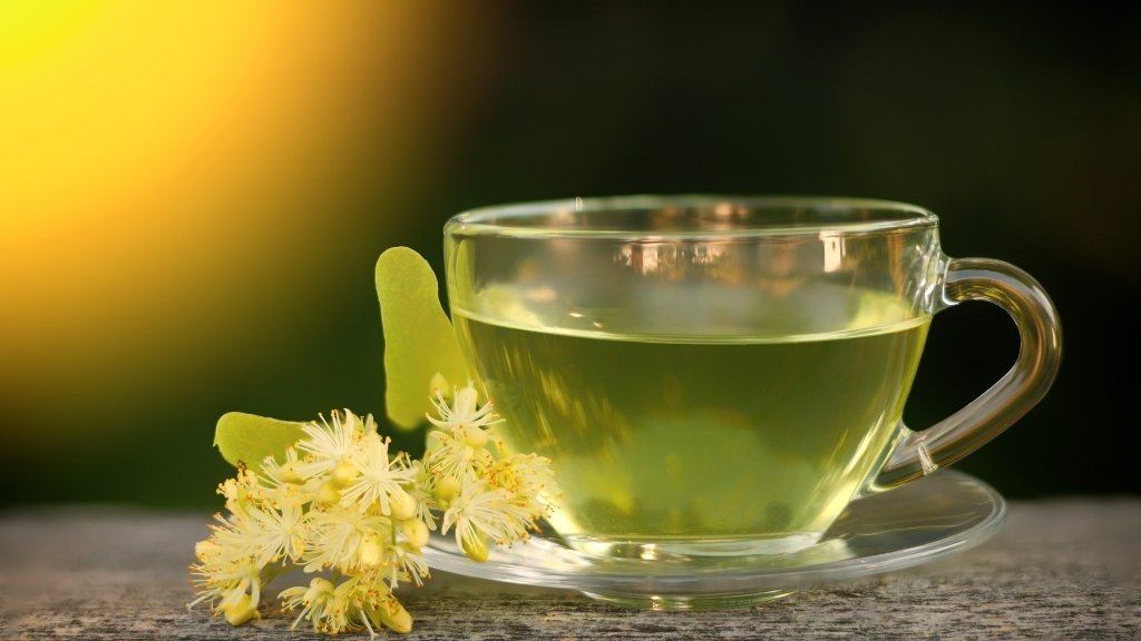 Linden flower tea in a clear glass cup with fresh linden blossoms on a wooden table.