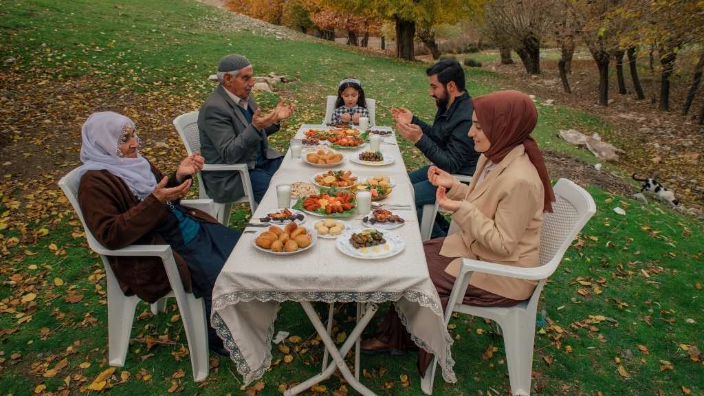 A traditional Turkish family gathered outdoors around a table, sharing food and praying together, reflecting cultural values of faith, unity, and hospitality.